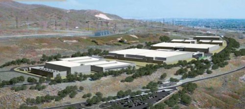 Aerial view of a large industrial complex surrounded by hills and power lines, with parking and greenery nearby.