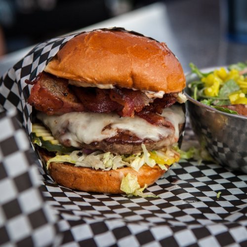 A juicy burger with bacon, melted cheese, and lettuce, served in a checkered paper wrapper alongside a salad.