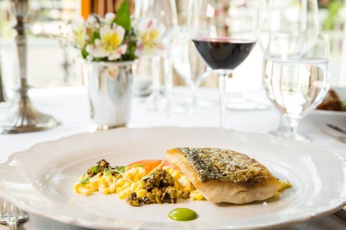 A beautifully plated dish featuring fish, pasta, and vegetables, with wine and flowers in the background.