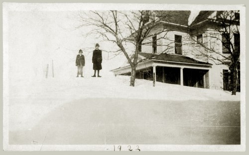 Two children stand in deep snow near a house, with trees bare of leaves, in a vintage photo from 1923.