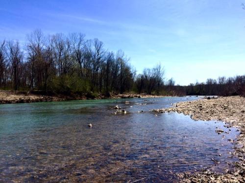 A serene river flows through a landscape with bare trees and rocky banks under a clear blue sky.