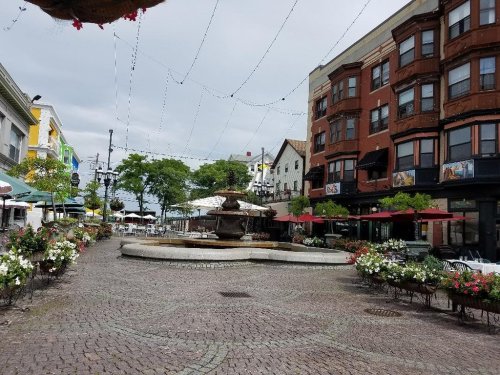 A quiet plaza with a fountain, surrounded by flower beds and outdoor seating at cafes, under a cloudy sky.