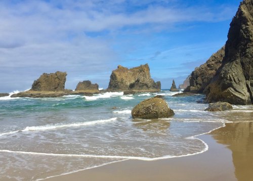 A scenic beach with rocky formations, gentle waves, and a blue sky with clouds.