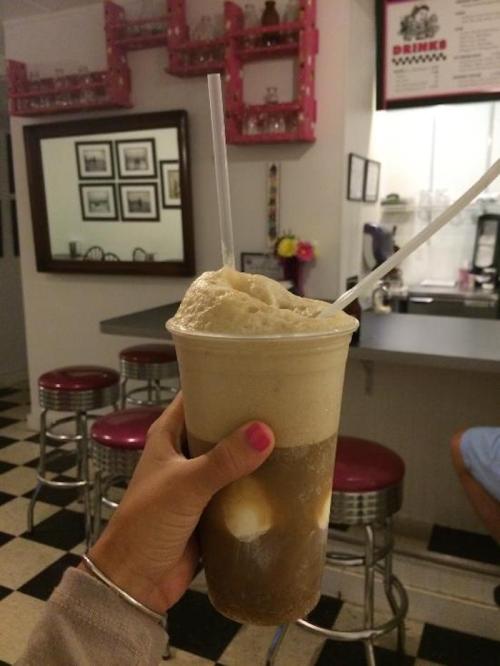 A hand holding a large frothy drink with two straws, set against a retro diner background.