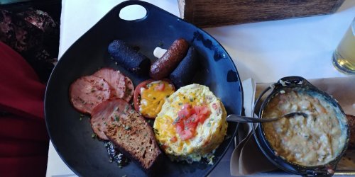 A black plate with an omelette, sliced meats, sausage, and a bowl of creamy porridge.