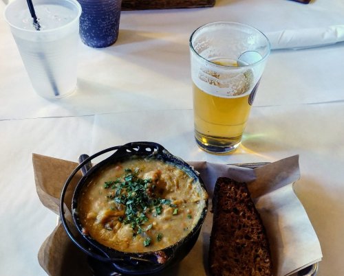 A bowl of creamy soup topped with herbs, served with a slice of bread and a glass of beer on a table.
