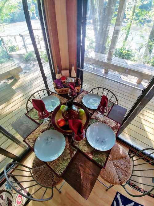 A beautifully set dining table with plates, glasses, and fruit, surrounded by large windows overlooking nature.
