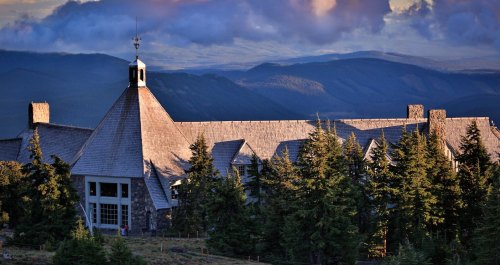 A mountain lodge with a peaked roof surrounded by trees, set against a backdrop of rolling hills and clouds.