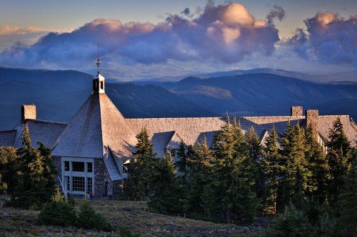 A stone building with a peaked roof surrounded by trees, set against a backdrop of mountains and clouds at sunset.