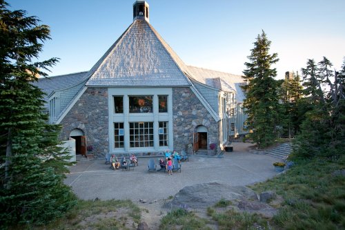 A large stone building with a triangular roof, surrounded by trees and people sitting outside on a patio.