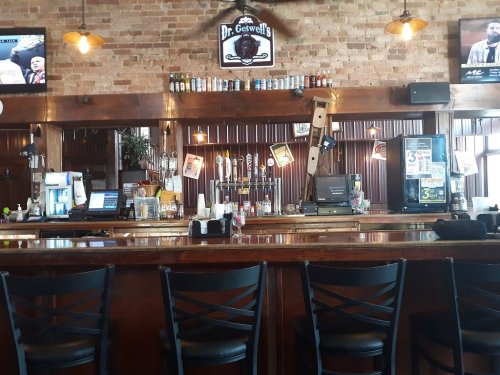 A wooden bar with stools, beer taps, and a brick wall backdrop, featuring various drinks and a menu display.