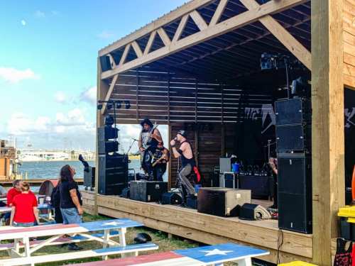 A band performs on an outdoor stage by the water, with colorful picnic tables in the foreground and a clear sky.