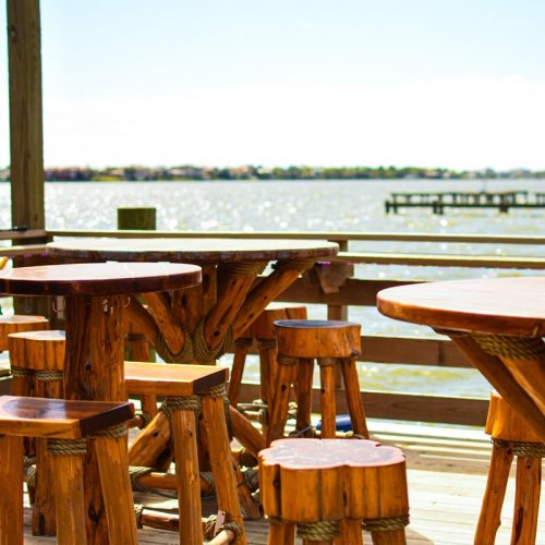 Wooden tables and stools on a deck overlooking a shimmering body of water under a clear blue sky.