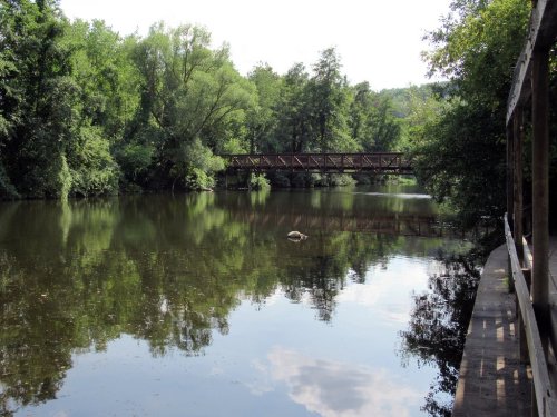 A serene river scene with lush greenery, a wooden bridge, and reflections on the water's surface.