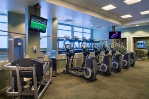 A modern gym with cardio machines, large windows, and two TVs displaying sports.