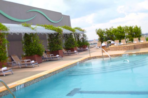 A serene rooftop pool area with lounge chairs, shaded by canopies and surrounded by greenery under a cloudy sky.