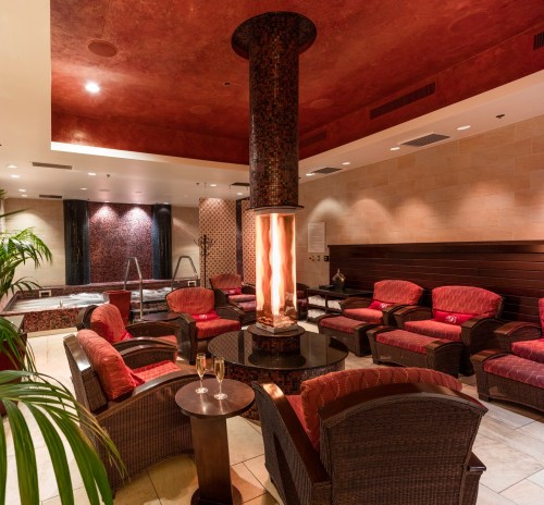 Cozy spa lounge with a central fireplace, plush seating, and a relaxing atmosphere, featuring a hot tub in the background.