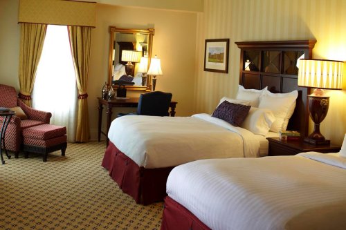 A cozy hotel room featuring two beds, a chair, a desk, and a window with curtains, all in warm tones.
