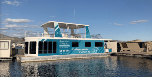 A houseboat with a blue and white design, advertising boat rentals, docked in a marina under a clear sky.