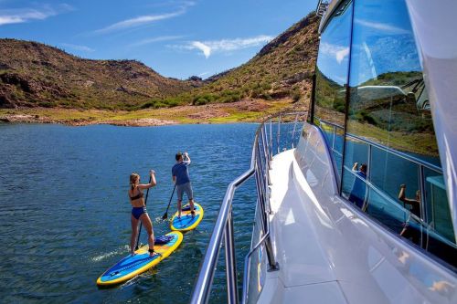 Two people paddleboarding on a calm lake surrounded by hills, with a boat in the foreground reflecting the scenery.