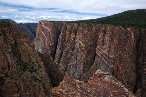 A dramatic view of steep, rocky cliffs with layered textures and greenery at the top under a cloudy sky.