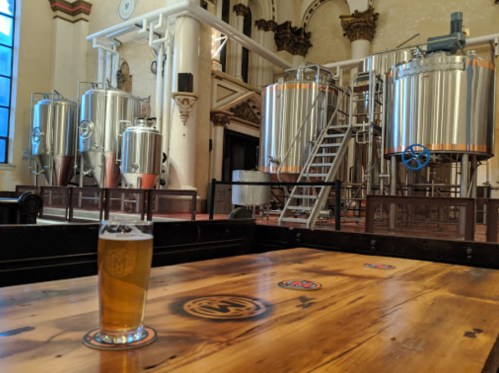 A glass of beer on a wooden table in a brewery with large brewing tanks in the background.