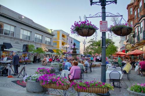 A lively outdoor plaza with diners, colorful flowers, and a fountain, set against charming buildings at dusk.