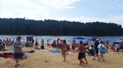 Crowded beach scene with people enjoying the water, sunbathing, and relaxing under umbrellas by a lake.