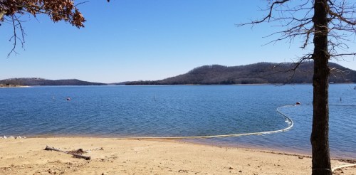 A serene lake view with clear blue water, sandy shore, and distant hills under a bright blue sky.