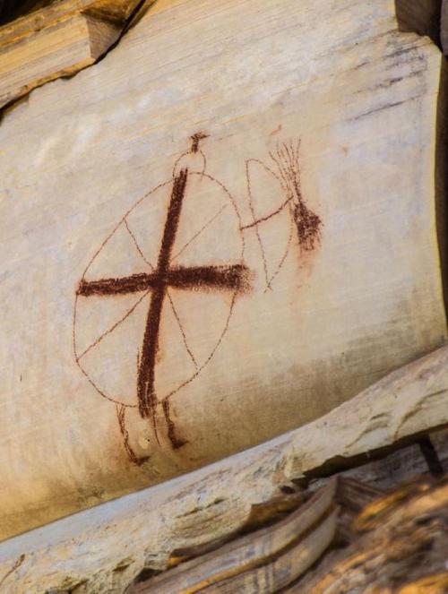Ancient rock art depicting a figure with a circular shield and a cross symbol, etched into a stone surface.