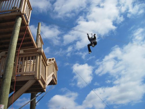 A person zip-lining from a wooden platform against a blue sky with clouds.