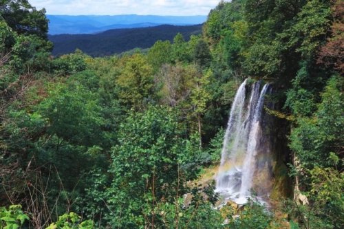 A scenic waterfall cascading down rocky cliffs, surrounded by lush green trees and distant mountains under a blue sky.