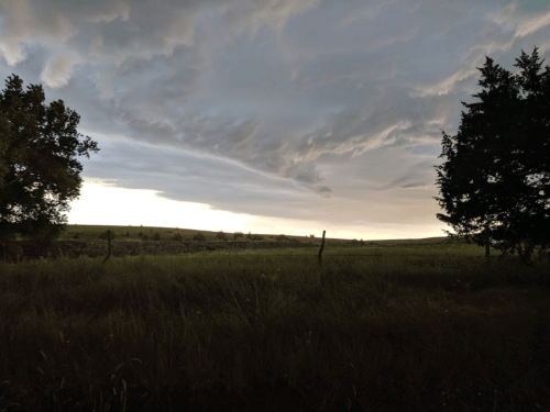 A dramatic sky with dark clouds over a grassy field, framed by trees on either side.