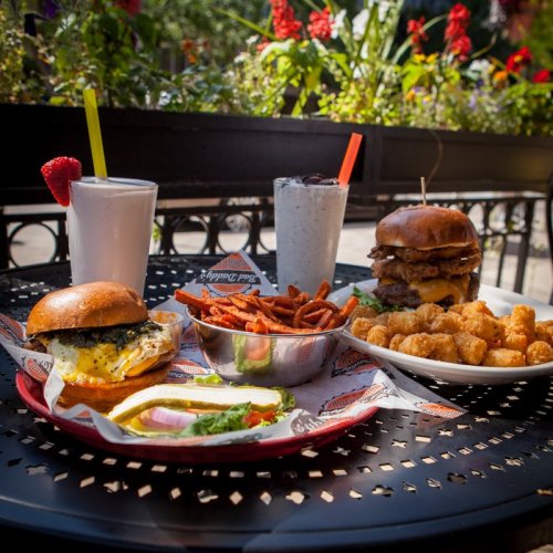 A table with a burger, sweet potato fries, tater tots, and milkshakes, surrounded by flowers in a sunny outdoor setting.