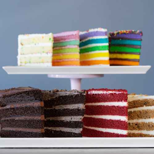 A display of colorful layered cakes on a white stand, featuring chocolate, red velvet, and rainbow varieties.