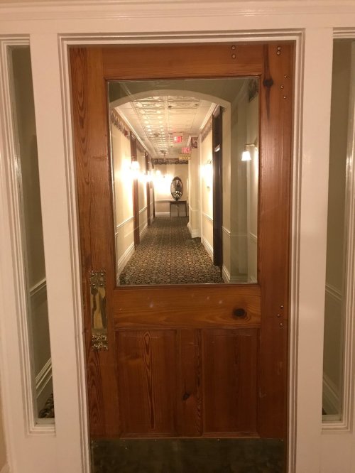 A wooden door with a glass panel opens to a well-lit hallway featuring patterned carpet and a mirror at the end.
