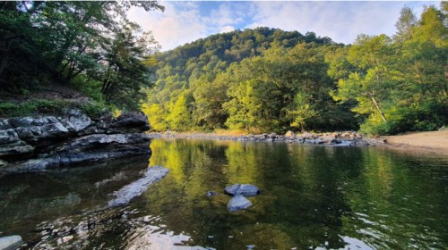 A serene river scene with lush green trees and a rocky shore, reflecting the blue sky and surrounding hills.