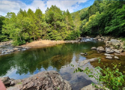 A serene river surrounded by lush green trees and rocky banks under a partly cloudy sky.