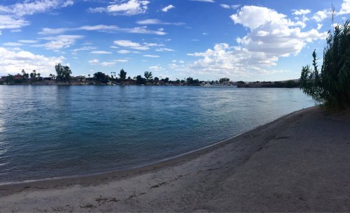 A serene river scene with clear blue water, sandy shore, and a backdrop of trees and clouds under a bright sky.