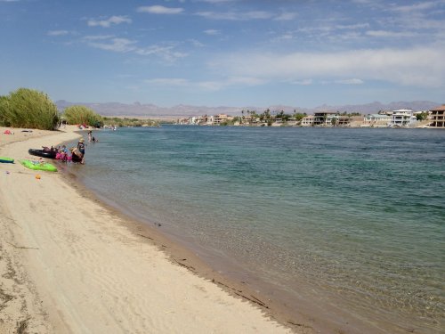 A sandy beach along a calm river, with people enjoying the water and houses visible in the background under a blue sky.