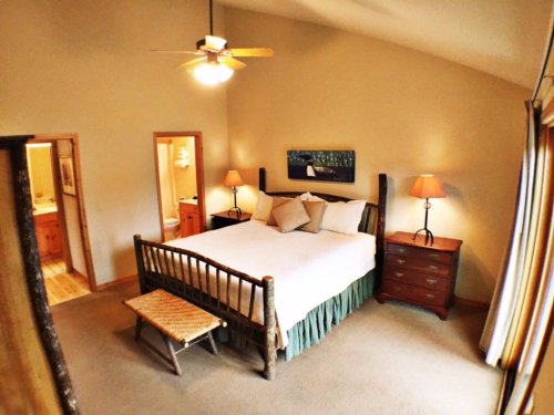 Cozy bedroom with a wooden bed, nightstands, lamps, and a ceiling fan, featuring warm tones and a serene atmosphere.