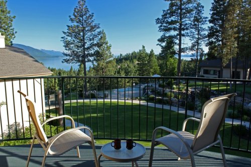 A balcony view overlooking a serene lake surrounded by trees, with two chairs and a small table in the foreground.