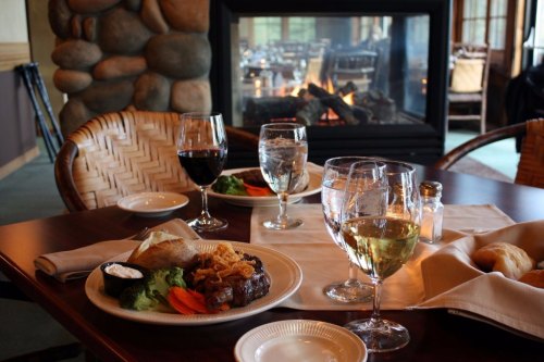 A cozy dining table with a steak dinner, vegetables, and drinks, set near a warm fireplace.