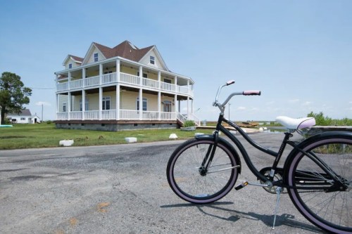 A black bicycle stands on a road in front of a large, multi-story house with a wraparound porch.