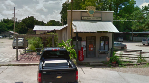 A small, rustic building with a sign, surrounded by greenery and a parked black truck in front.