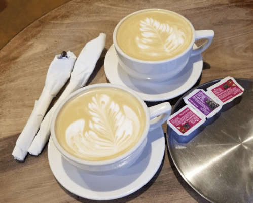 Two cups of latte with leaf patterns on top, accompanied by sugar packets and napkins on a wooden table.