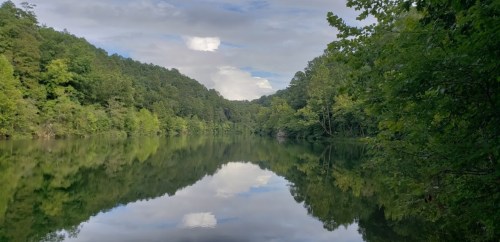 A serene lake surrounded by lush green trees, reflecting the sky and mountains in calm waters.