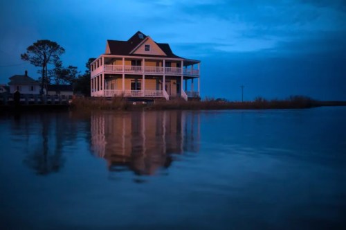 A large house by the water at dusk, reflecting in the calm surface, surrounded by trees and a cloudy sky.