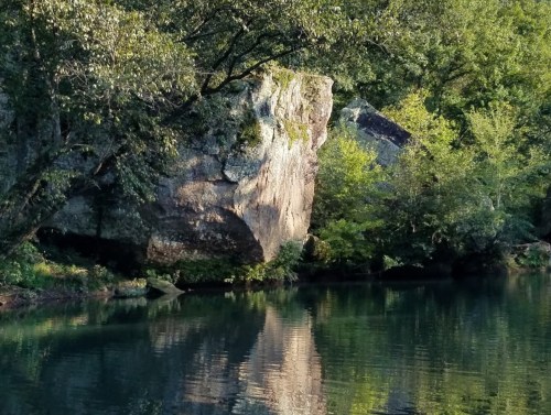 A serene river scene featuring a large rock and lush greenery reflected in calm water.