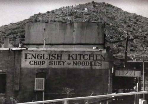 Black and white photo of a building with a sign reading "English Kitchen Chop Suey Noodles" near a mountain.